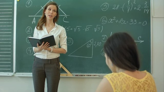 Teacher or docent or educator giving while lesson in front of a blackboard or board a sheet of paper and educate or teaching students or pupils or mates in a school or class