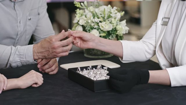 Closeup of hands of shop assistant giving silver wedding ring to man and then he putting it on finger of his bride in jewelry salon