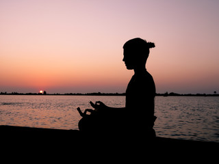 A young boy sits at a riverbank and meditates at sunset.