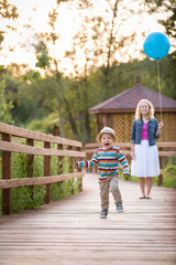 Fototapeta na wymiar Beautiful young woman walking on wooden bridge, holding big blue balloon and looking at her little cute running toddler boy on the sunset in the park. Birthday. Mother and son. Happy family..
