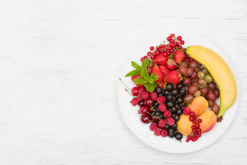 Assortment of juicy fruits on white plate and white table background. Banana, strawberries, blueberries, raspberries, gooseberry, blackcurrant decorated with mint for summer dessert or snack.