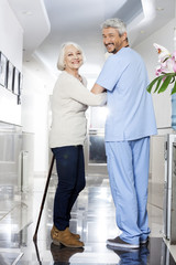 Senior Woman With Stick And Physiotherapist Standing In Rehab Ce
