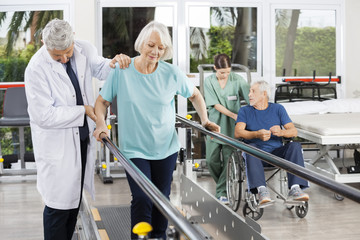 Doctor Helping Woman To Walk Between Bars In Fitness Center