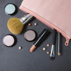 A pastel pink make up bag with cosmetic products spilling on a dark slate background