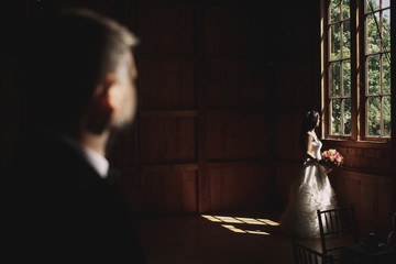 Groom admires a dreamy bride standing before the window full of