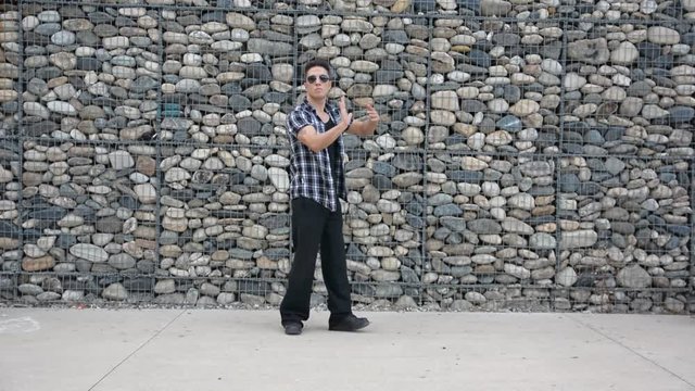 Young male trendy hip-hop dancer outside, doing break-dance in city setting in front of stone wall