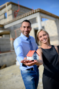 business man and woman realtor holding a model house in front of building construction site 