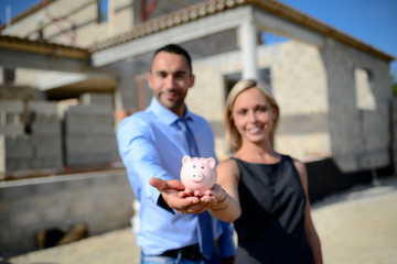 business man and woman realtor holding piggy bank symbol in front of house building construction site