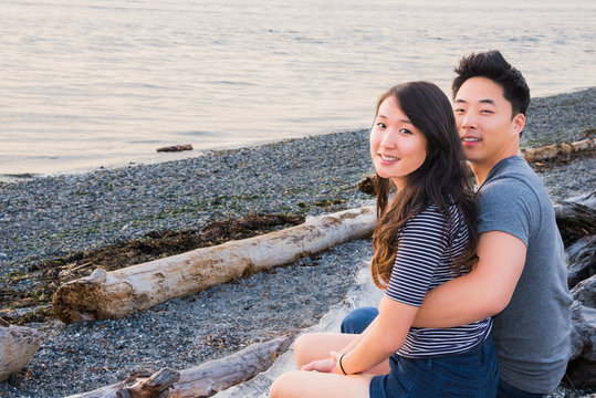 Young Asian Couple Sitting on Driftwood on Beach. Copy space.