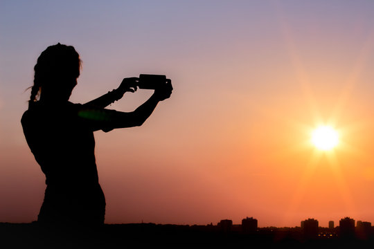 Silhouette of female taking photos of sunset with smart phone. Summer, travel and technology concepts.