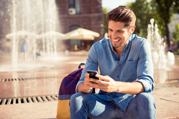 cheerful young man sitting near  fountain and looking phone