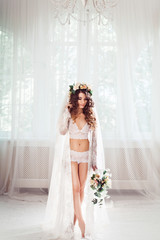 sexy bride standing on a white light hall dressed in nighty, bridal veil and wreath, with bouquet...