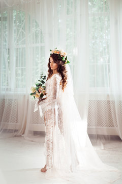 sexy bride standing on a white light hall dressed in nighty, bridal veil and wreath, with bouquet of flowers