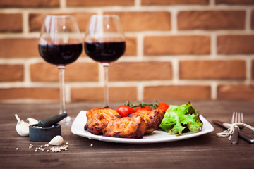 Juicy pieces of grilled meat fillet served with cherry tomatoes branch and lettuce on a white plate, glass of red wine, knife, fork, on the brick wall background. Romantic concept. Horizontal