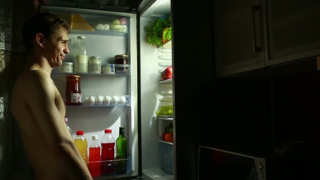 night man stands at the open refrigerator. eating sandwich. hungry guy