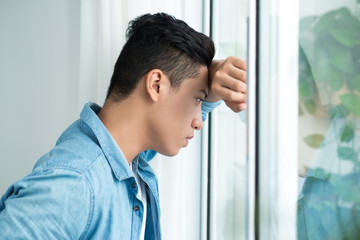 Thoughtful asian man looking out the window in bedroom at home