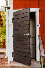 Old weathered and black toilet door made of wood at the corner of a red and white wooden building.