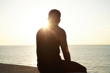 Silhouette of pensive African young male sitting on sidewalks at seaside watching sun setting over sea after outdoor training, waiting for his girlfriend, thinking about his relationships. Flare sun