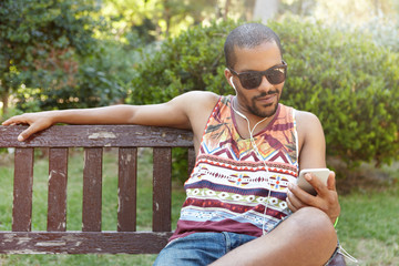 African guy in earphones sitting on bench in city park listening to music on his smartphone, checking e-mail using Internet-enabled mobile phone, liking posts and leaving comments on social networks