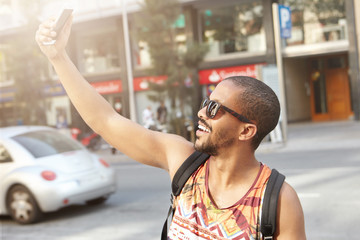 Fototapeta premium Stylish dark-skinned college student with backpack on his shoulders taking selfie using mobile phone posing and smiling, standing in middle of street on his way to university. Modern lifestyle concept