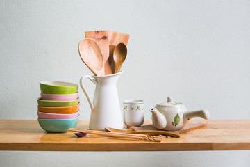 Kitchenware with wooden ladle and wooden spoon in white vase, set of tea cup and set of bowl on wooden table over white wall background