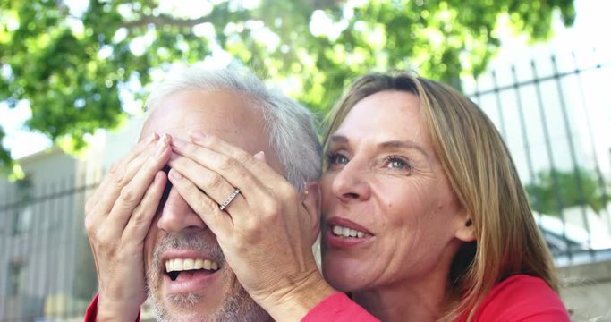 Mature woman is hiding her husband's eyes in the street on a sunny day 