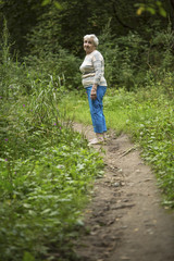 An elderly woman stands on a footpath in a green forest.
