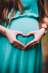 Young pregnant woman standing near the tree in the turquoise green dress. Hands on his stomach in the shape of heart