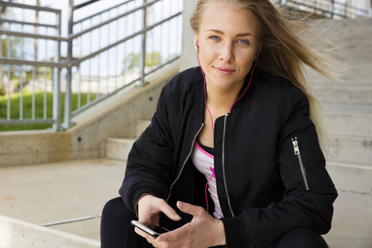 Smiling blonde woman sits outdoor and uses phone