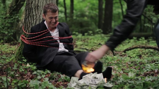 Businessman tied to a tree frantically tries to to put out burning dollars.