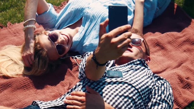 Young couple having fun relaxing together in the sun lying on their backs on a rug on the grass in their sunglasses chatting and smiling