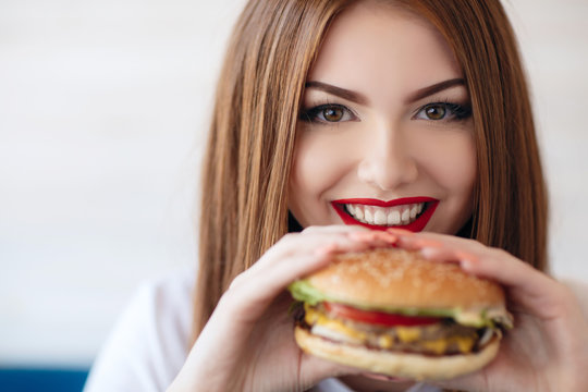 Beautiful woman with red long straight hair,bright make-up,brown eyes,red lipstick,long eyelashes,pink nail polish in the hands holding a large hamburger, sitting at a table in a cafe,dinner alone