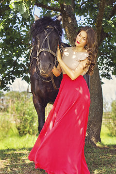 Young european woman in a long red dress and black beautiful horse standing near a big oak tree