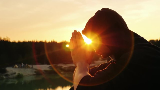 Young man praying at sunset. He sits in profile to the camera hood, clasped his hands together in prayer
