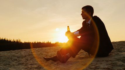 Young businessman in a suit sitting in the sand and drink alcohol from a flask