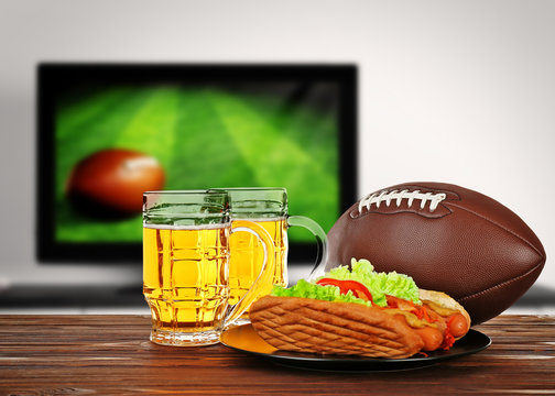 Two glasses of beer, ball and snack on wooden table in front of television show of football. Watching football match at home.