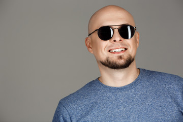 Portrait of confident handsome middle-aged man in grey shirt and sunglasses posing smiling at camera over dark background.