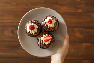 Female hand holding plate with cupcakes on wooden background, top view