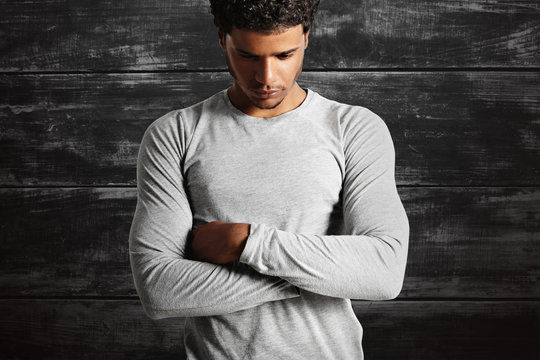 Thoughtful-looking fit dark-skinned model wearing a plain cotton light gray t-shirt with his arms crossed on the background of a black painted wooden wall.