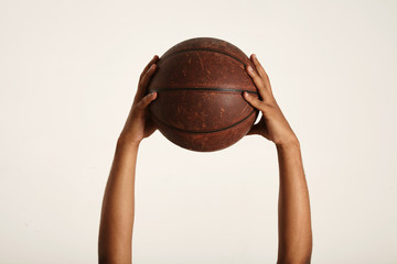 Close up shot of two strong dark-skinned hands holding up an old vintage leather brown basketball, isolated on white
