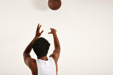 Back shot of a muscular fit black player in white sleeveless shirt throwing a brown leather basketball away from camera isolated on white