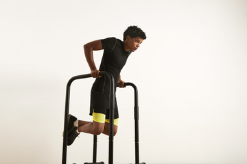 Fototapeta na wymiar A portrait of a focused muscular African American young man in black workout clothes doing dips on parallel bars against white background