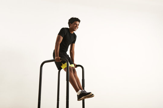 Low angle shot of a smiling strong African American athlete with an afro wearing black synthetic shirt and black and yellow shorts doing L-sits on short bars at home isolated on white.