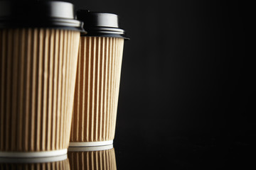 Two identical craft cardboard coffee cups with black lids on reflective table against black wall on the side of a horizontal shot