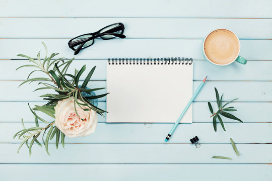 Morning Coffee Cup, Clean Notebook, Pencil, Eyeglasses And Vintage Rose Flower In Vase On Blue Rustic Desk Overhead View. Planning And Design Concept. Cozy Breakfast. Flat Lay Styling.