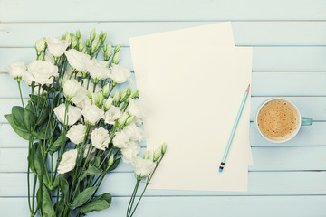Morning coffee cup, empty paper list, pencil, and bouquet of white flowers eustoma on blue rustic table from above. Woman working desk. Cozy breakfast. Flat lay styling.