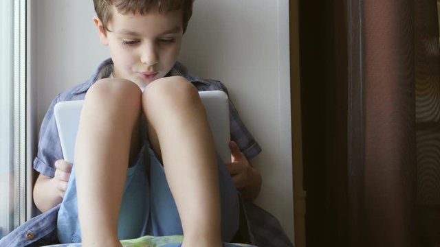 A cute little boy uses a white tablet PC on a windowsill at home