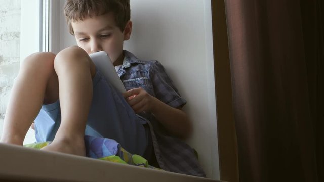 BOTTOM VIEW: A cute little boy uses a white tablet PC on a windowsill at home