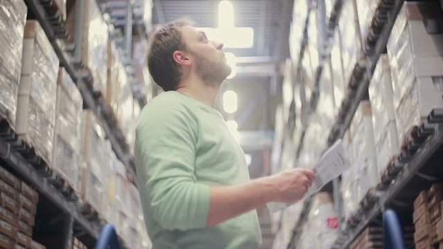 A man is standing near shelves with goods, checking his list, looking for necessary stuff in a warehouse