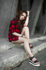 Fototapeta na wymiar Beautiful young hipster girl posing and smiling near urban wall background in red plaid shirt, shorts, outdoors summer portrait.